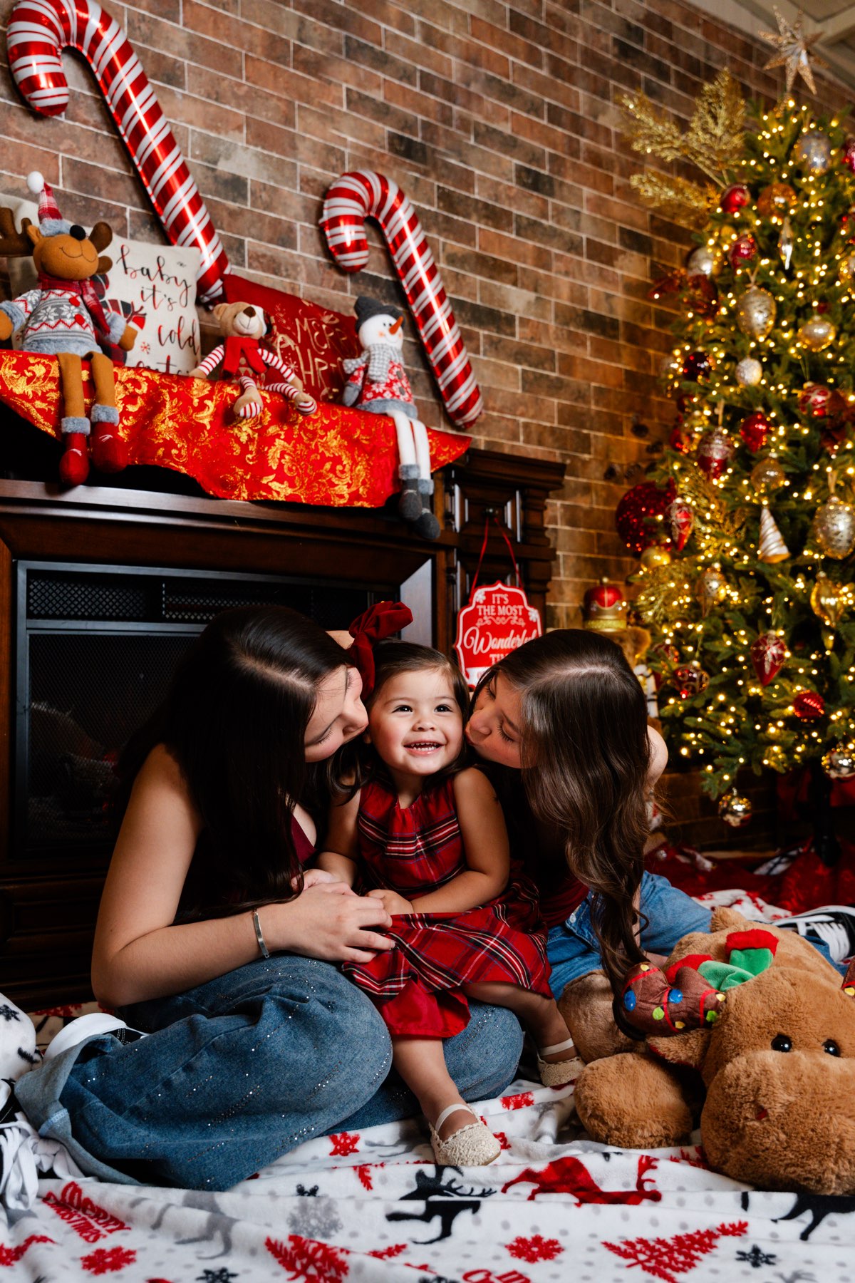 Dos hermanas dándole un beso a la más chica, que se ríe, frente a la chimenea del set navideño