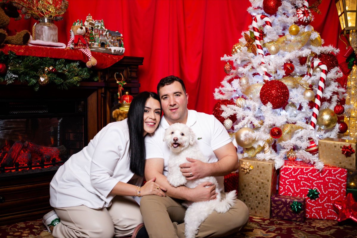 Una pareja vestida de blanco abrazando a su perro junto al árbol blanco y los regalos