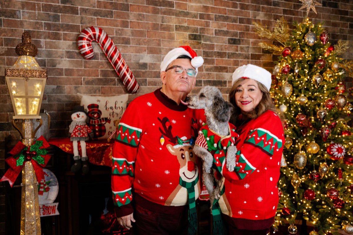 Una pareja con suéteres navideños y su perro, que le da un beso a él