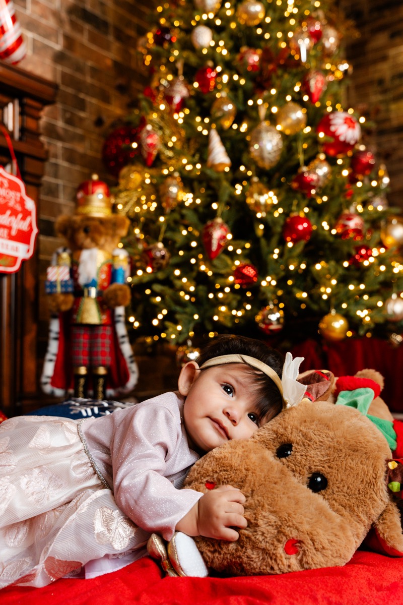 Una bebé acostada sobre el reno de peluche, con el árbol iluminado detrás