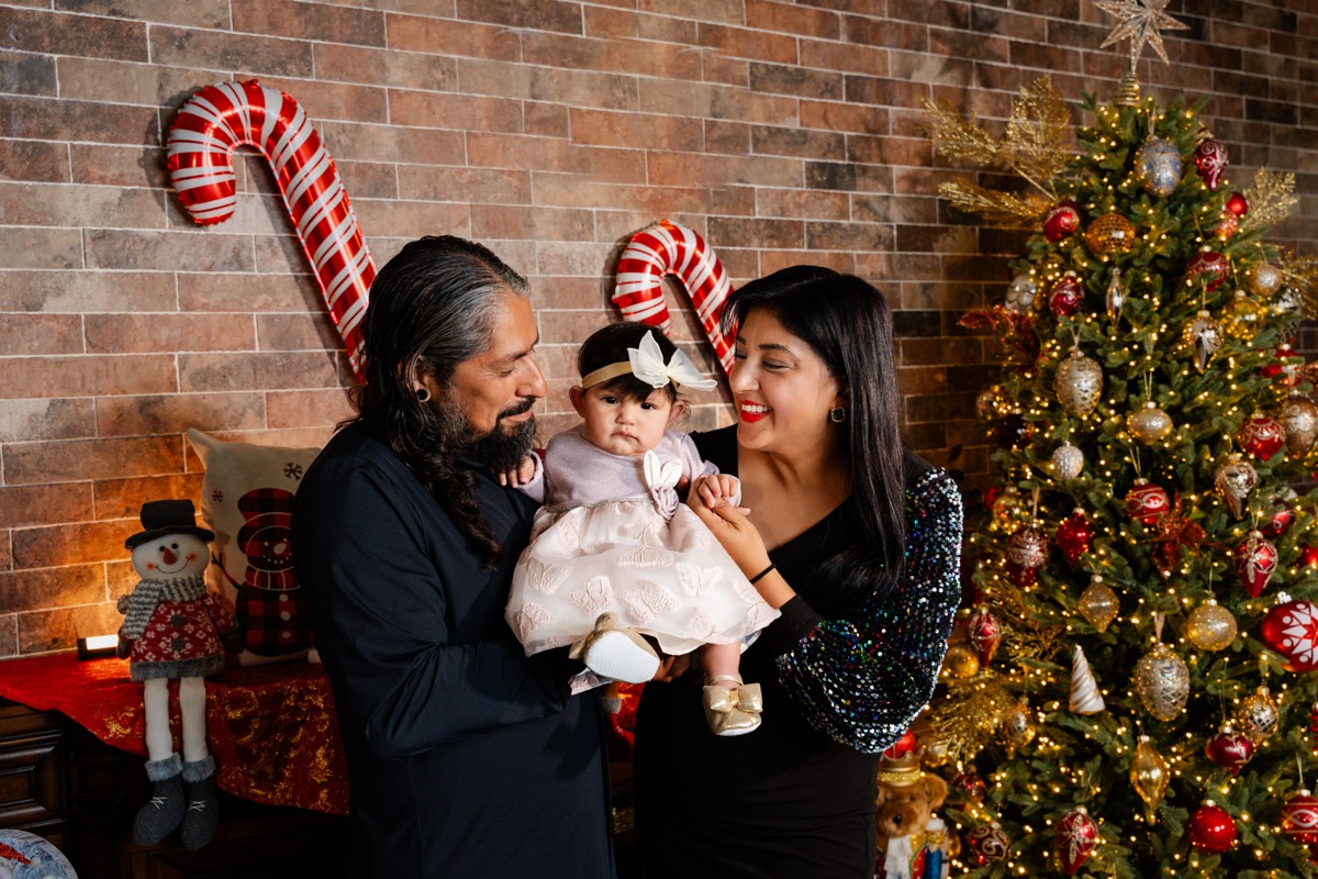 Papá y mamá mirando a su bebé de vestido rosa, junto al árbol